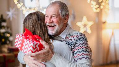 Portrait of young woman giving present to happy grandfather indoors at home at Christmas.