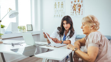 A female doctor sits at her desk and chats to an elderly female patient while looking at her test results. Patient Having Consultation With Female Doctor In Office