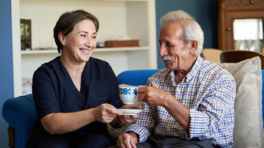Eine Frau reicht ihrem pflegebedürftigen Vater eine Tasse Kaffee.