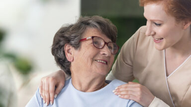 Eine junge Frau beugt sich über eine ältere und lächelt sie an.