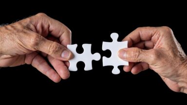 Man sieht rechts und links je eine Hand einer älteren Person mit einem Puzzleteil in der Hand