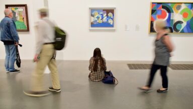 Das Bild zeigt Menschen in einem Museum vor drei Bildern. Eine der Personen sitzt auf dem Boden und betrachtet das Bild in der Mitte.