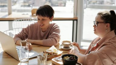 Zwei Frauen sitzen am Tisch und schauen auf den Laptop. Eine Frau hat das Down Syndrom.