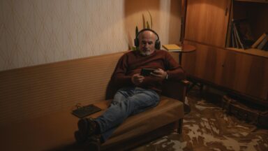 Man sieht einen älteren Mann auf einem Sofa sitzend mit Kopfhörern auf den Ohren