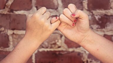 Zwei Hände deren kleine Finger ineinander verhakt sind. Im Hintergrund eine rote Ziegelsteinmauer.