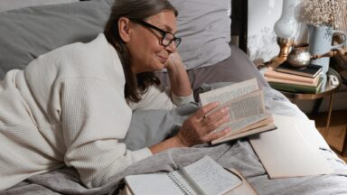 Man sieht eine ältere Frau auf einem Bett auf dem Bauch liegend und in einem Buch blättern